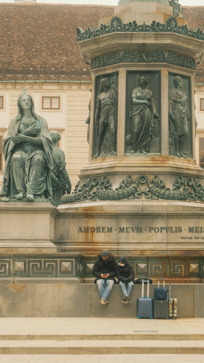 Two travelers seated by a historic monument with luggage in Vienna.